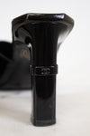 A close-up of a black patent leather high-heeled shoe heel, showing a Chanel logo band around the middle, on a white surface.
