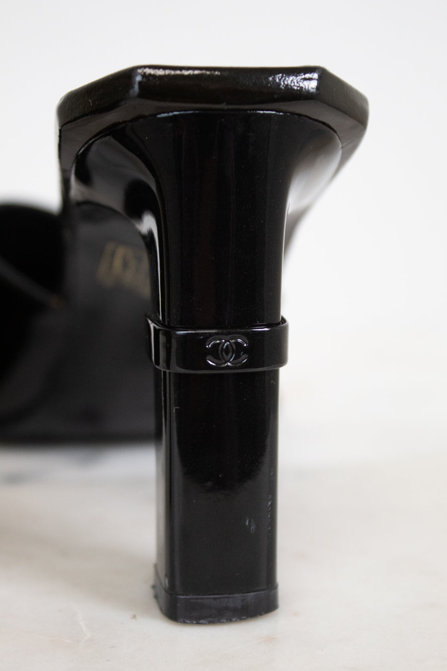 A close-up of a black patent leather high-heeled shoe heel, showing a Chanel logo band around the middle, on a white surface.