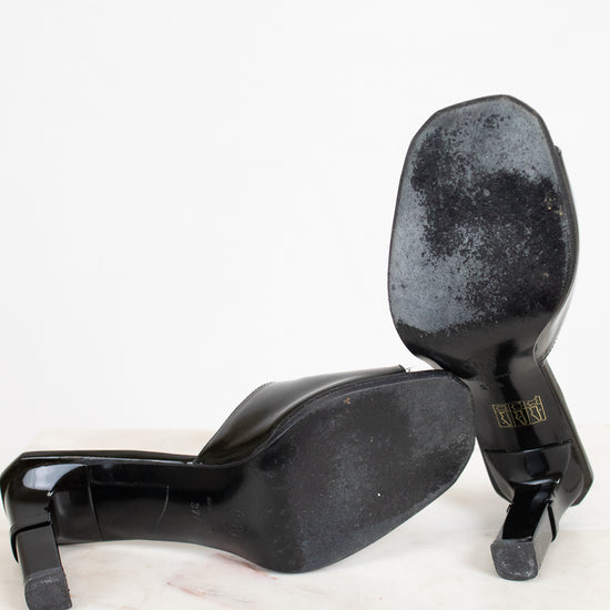 A pair of black patent leather high-heeled pumps shown from the sole, resting on a white marble surface against a white background.