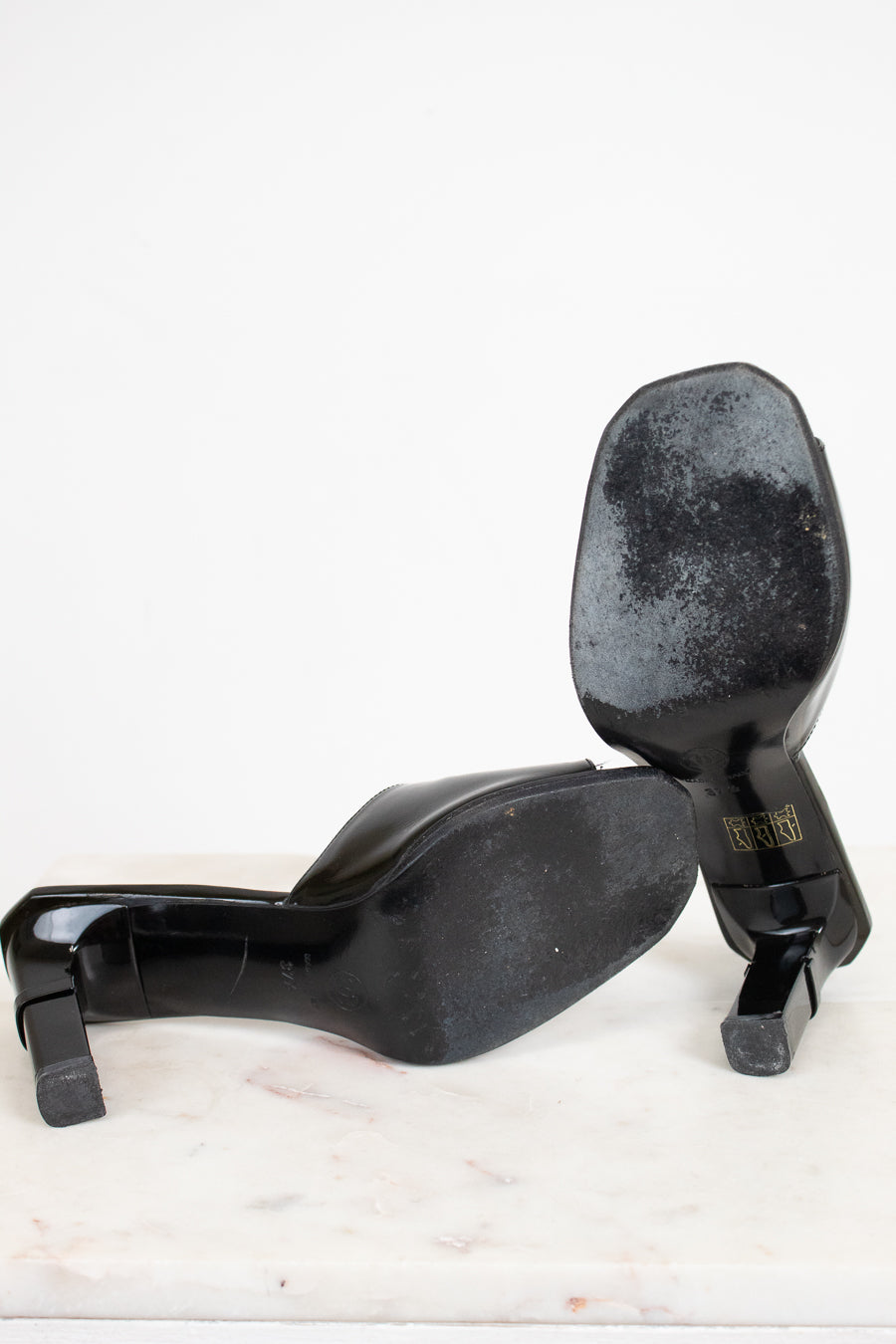 A pair of black patent leather high-heeled pumps shown from the sole, resting on a white marble surface against a white background.