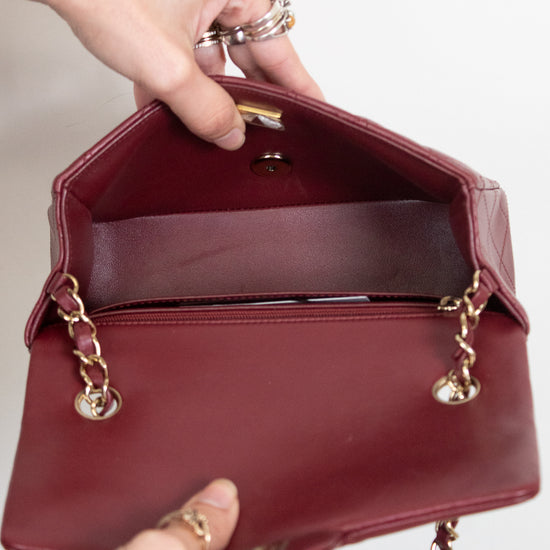 A burgundy leather handbag with gold-tone chain strap details, held open by two hands against a plain white background.