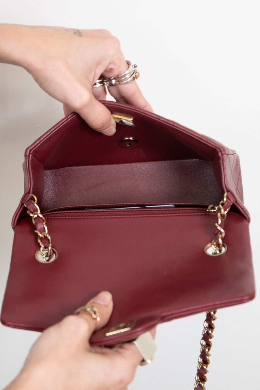 A burgundy leather handbag with gold-tone chain strap details, held open by two hands against a plain white background.
