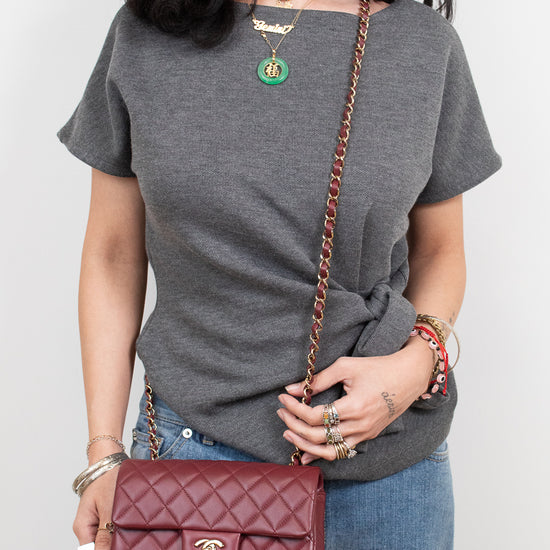A woman wearing a gray t-shirt and blue jeans holds a dark red quilted Chanel shoulder bag with a gold chain strap.