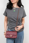 A woman wearing a gray t-shirt and blue jeans holds a dark red quilted Chanel shoulder bag with a gold chain strap.
