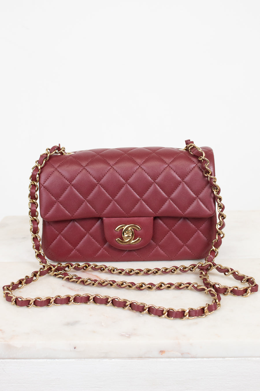 A classic quilted flap shoulder bag in burgundy leather with a gold-tone interlocking CC logo and a gold chain strap, displayed on a light marble surface against a white background.