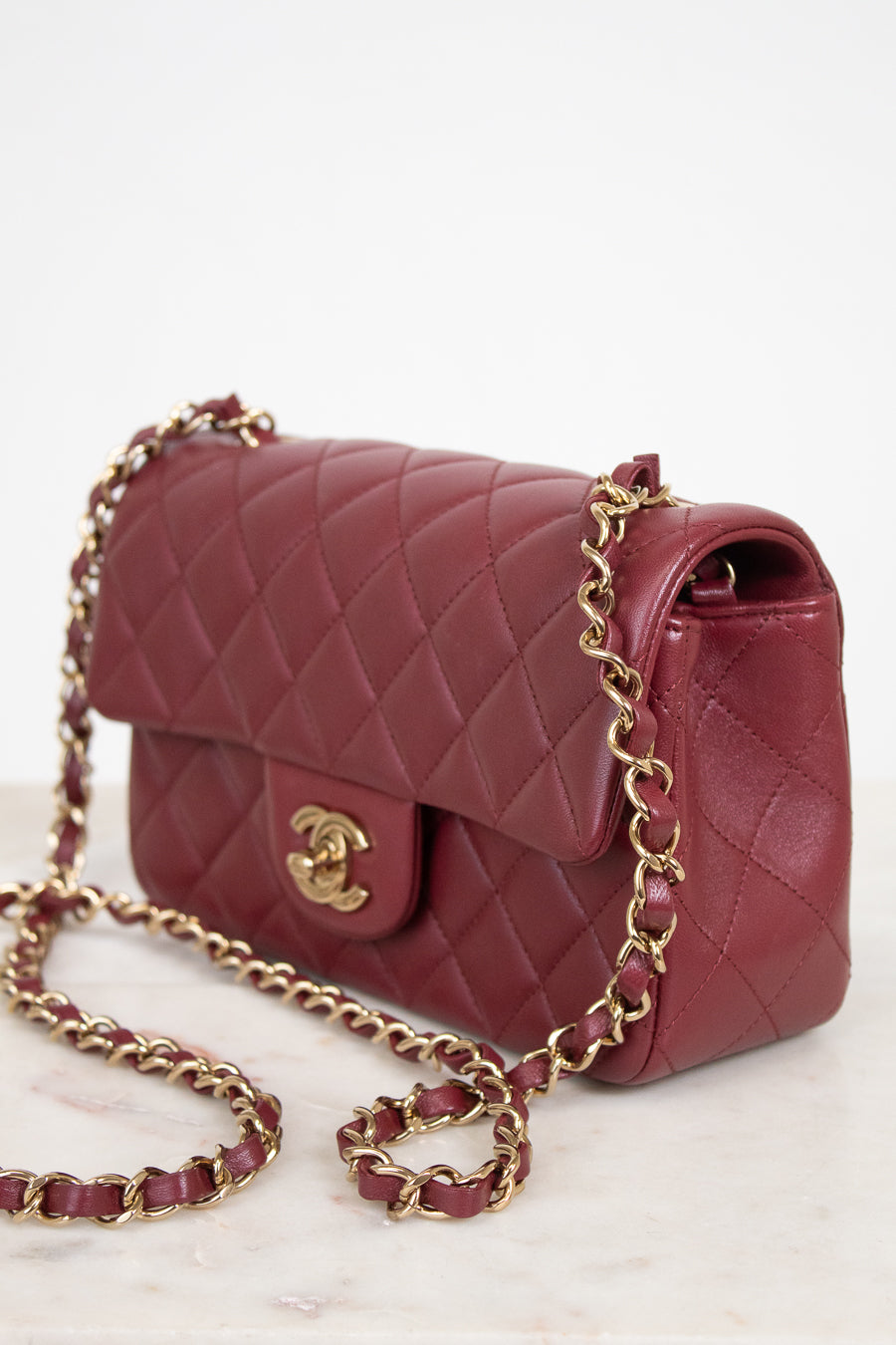 A quilted burgundy leather shoulder bag with a gold-tone chain strap and a gold interlocking CC logo, displayed on a light marble surface against a white background.