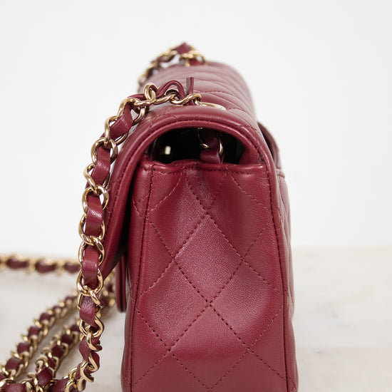 A close-up side view of a quilted burgundy leather handbag with a gold-tone chain strap, resting on a light marble surface against a white background.