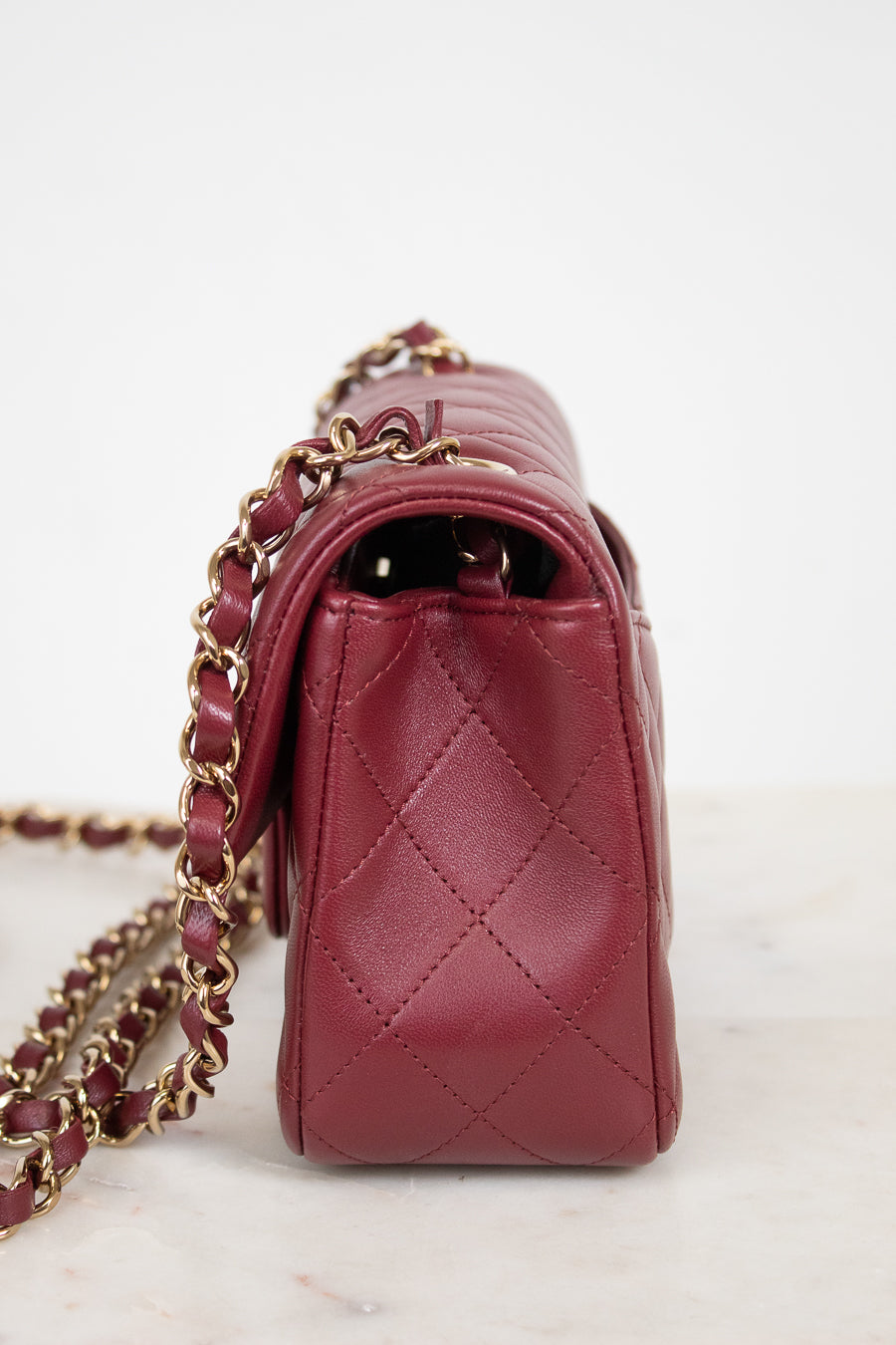 A close-up side view of a quilted burgundy leather handbag with a gold-tone chain strap, resting on a light marble surface against a white background.