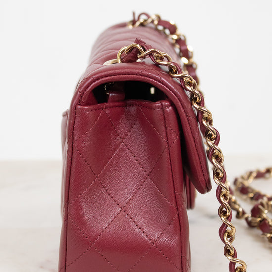 A close-up side view of a quilted burgundy leather handbag with a gold chain strap, resting on a light marble surface against a white background.
