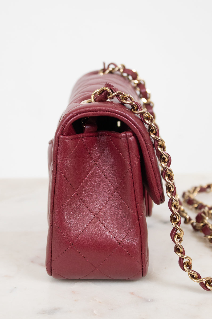 A close-up side view of a quilted burgundy leather handbag with a gold chain strap, resting on a light marble surface against a white background.