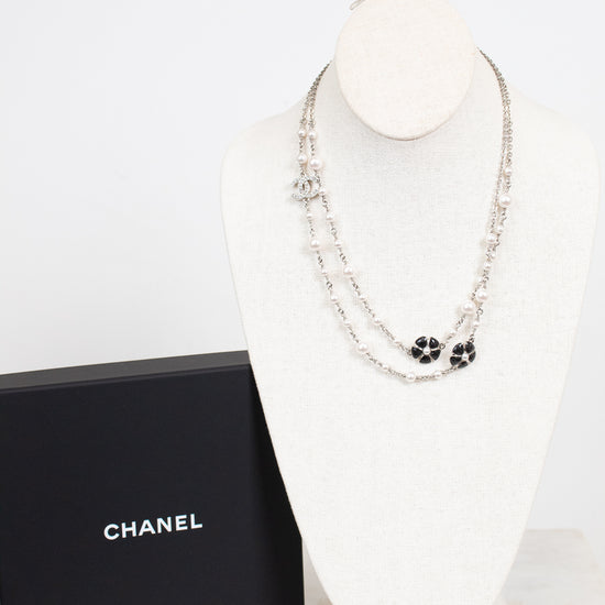 A Chanel necklace displayed on a white mannequin bust next to a black Chanel box. The necklace features silver-tone chains with black enamel floral clover motifs and silver-tone interlocking CC logos.