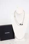 A Chanel necklace displayed on a white mannequin bust next to a black Chanel box. The necklace features silver-tone chains with black enamel floral clover motifs and silver-tone interlocking CC logos.