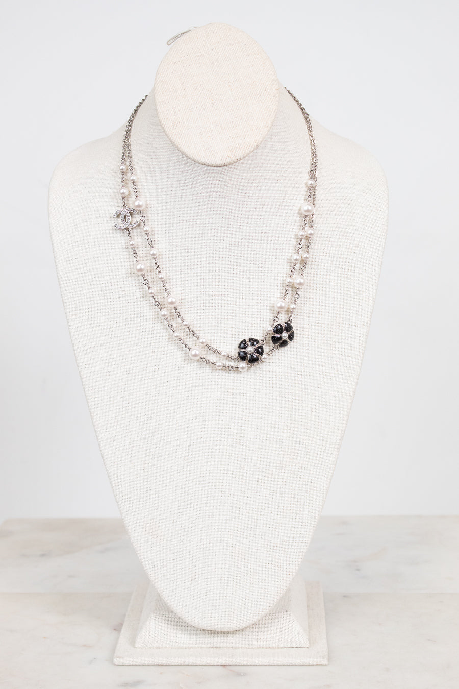 A double-strand silver-toned necklace displayed on a white fabric jewelry bust. The necklace features small silver beads, a silver chain with clear rhinestones, and two black floral cluster charms with clear rhinestones.