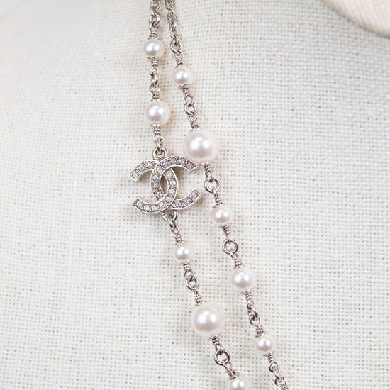 A Chanel double-strand pearl necklace with a silver-tone CC logo charm encrusted with clear crystals, displayed on a cream-colored fabric mannequin bust.