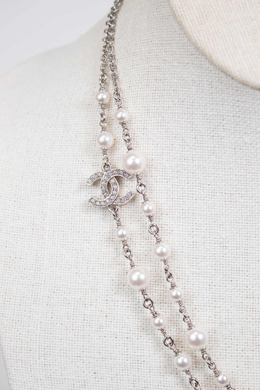A Chanel double-strand pearl necklace with a silver-tone CC logo charm encrusted with clear crystals, displayed on a cream-colored fabric mannequin bust.