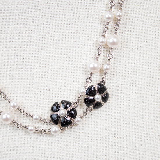 A double-strand necklace featuring white faux pearls and silver-tone chains, accented with black onyx flower motifs on a textured off-white fabric background.