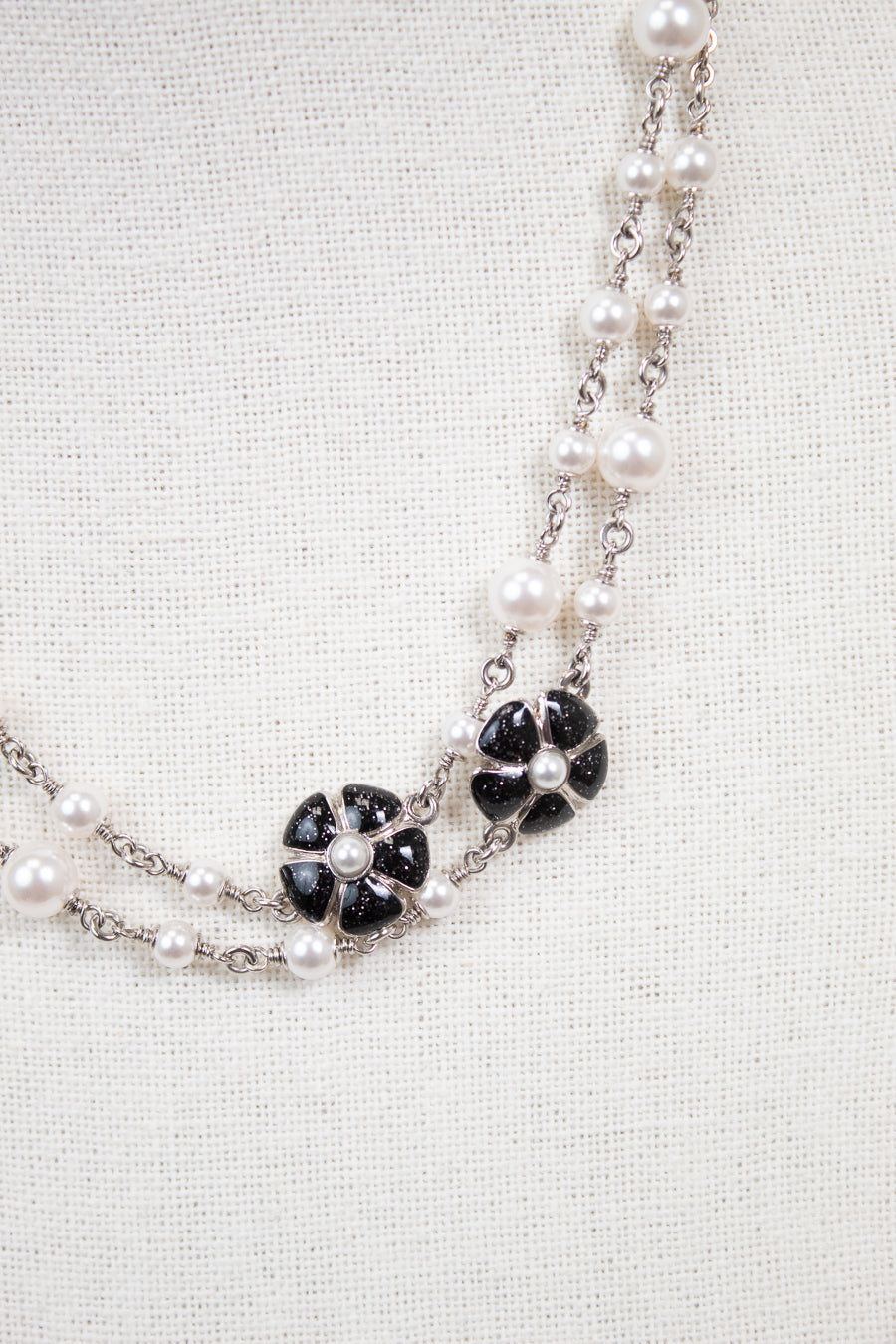 A double-strand necklace featuring white faux pearls and silver-tone chains, accented with black onyx flower motifs on a textured off-white fabric background.