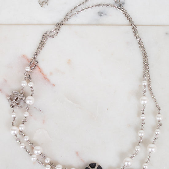 A double-strand silver chain necklace featuring white faux pearls and black floral accents, displayed on a light marble surface.