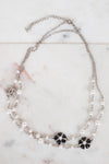 A double-strand silver chain necklace featuring white faux pearls and black floral accents, displayed on a light marble surface.