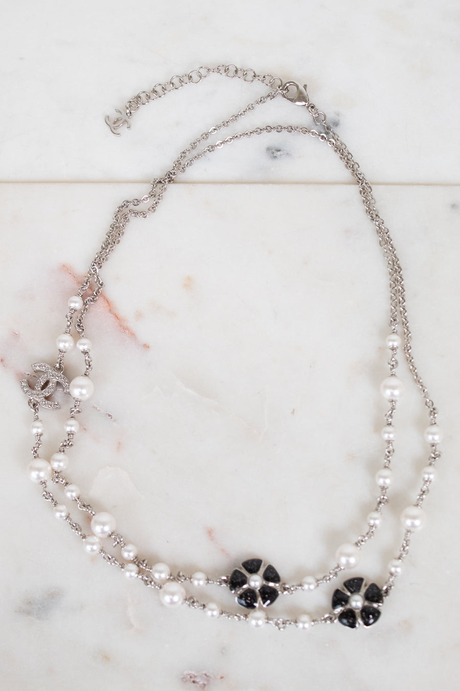 A double-strand silver chain necklace featuring white faux pearls and black floral accents, displayed on a light marble surface.