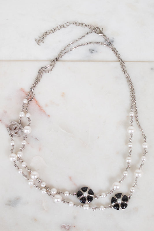 A double-strand silver chain necklace featuring white faux pearls and black floral accents, displayed on a light marble surface.