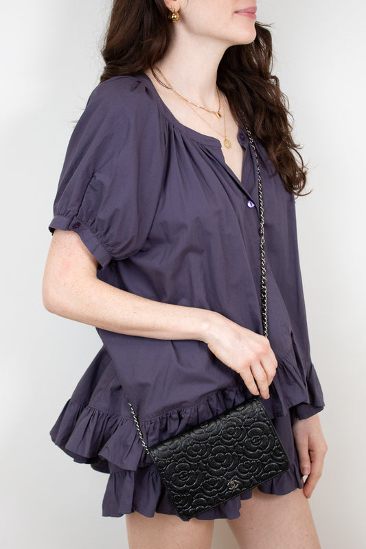 Woman wearing a purple blouse with ruffled details holding a black clutch with silver details against a white background