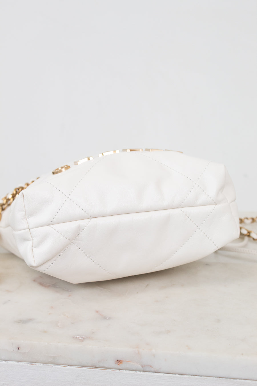 A white quilted leather shoulder bag with a gold chain strap, resting on a light marble surface against a white background.