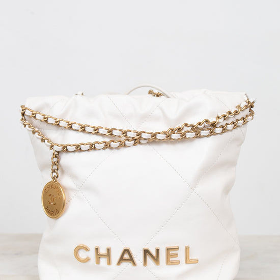A white quilted leather Chanel bucket bag with gold-tone chain detailing and a gold CHANEL logo on the front, set against a light gray background.