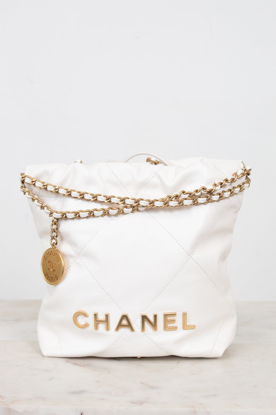 A white quilted leather Chanel bucket bag with gold-tone chain detailing and a gold CHANEL logo on the front, set against a light gray background.