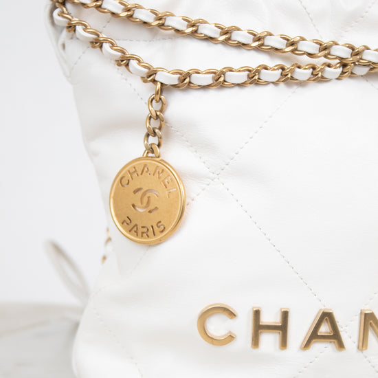 A close-up of a white quilted Chanel shoulder bag with gold-tone chain detailing and a round gold-tone hanging charm engraved with 'CHANEL PARIS'.