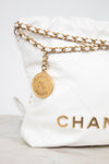 A close-up of a white quilted Chanel shoulder bag with gold-tone chain detailing and a round gold-tone hanging charm engraved with 'CHANEL PARIS'.