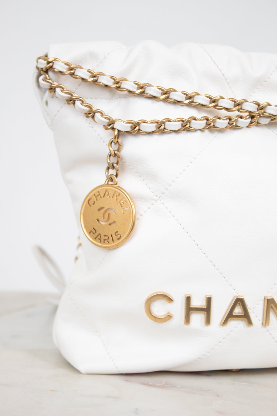 A close-up of a white quilted Chanel shoulder bag with gold-tone chain detailing and a round gold-tone hanging charm engraved with 'CHANEL PARIS'.