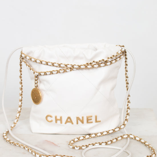 A white quilted Chanel shoulder bag with gold-tone chain straps and a gold CHANEL logo, displayed on a light marble surface against a white background.