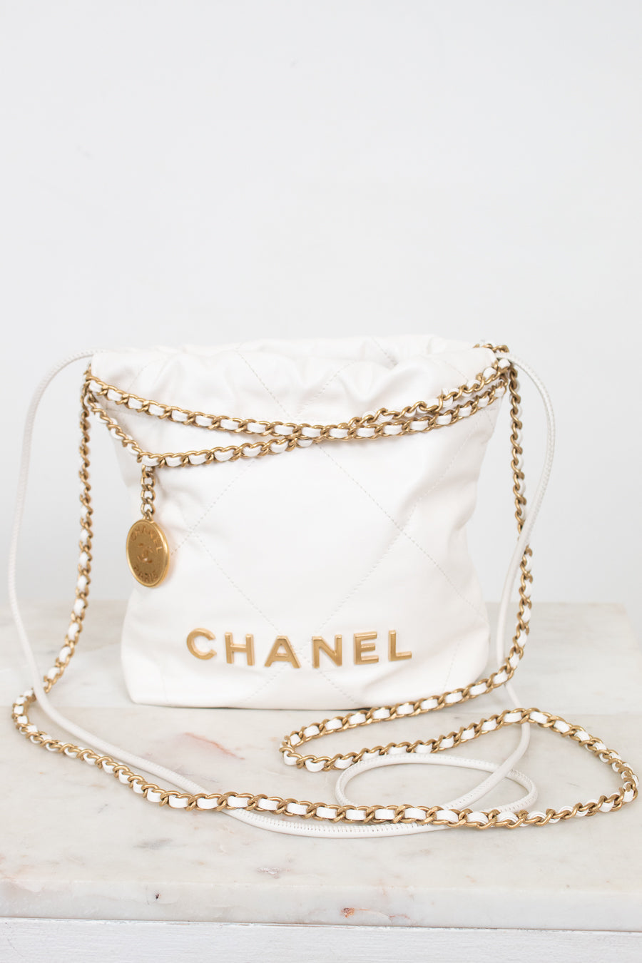 A white quilted Chanel shoulder bag with gold-tone chain straps and a gold CHANEL logo, displayed on a light marble surface against a white background.