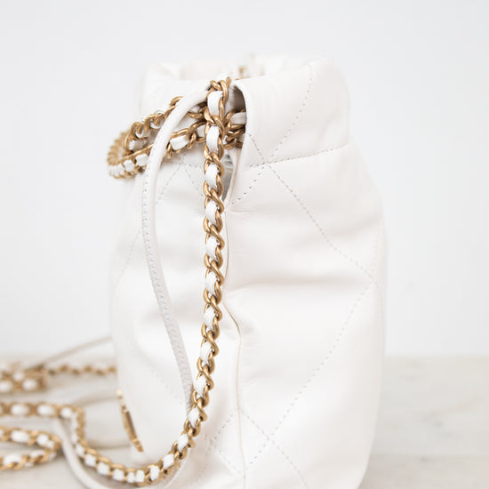 A white quilted leather bucket bag with a gold chain strap, resting on a light marble surface against a white background.