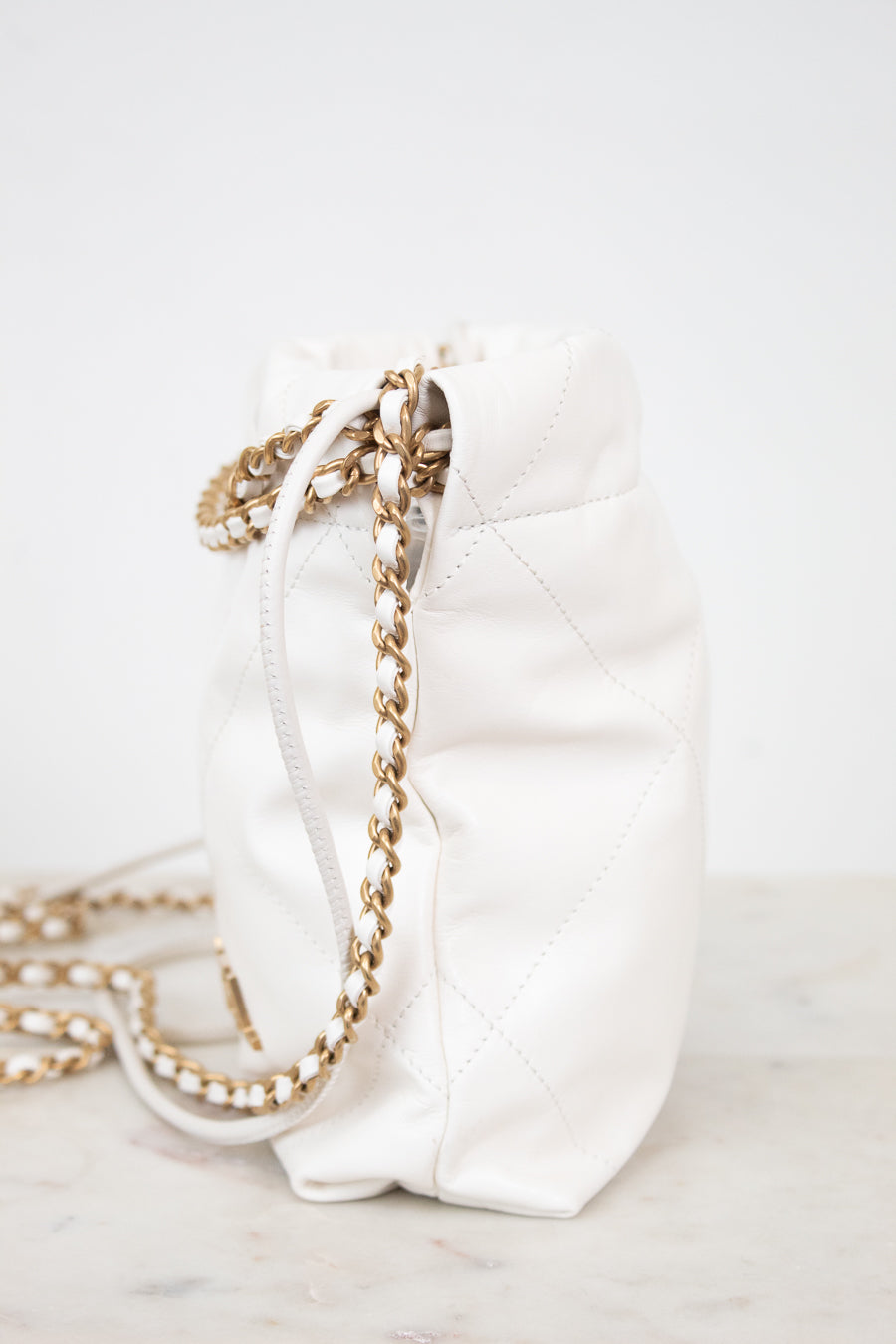 A white quilted leather bucket bag with a gold chain strap, resting on a light marble surface against a white background.