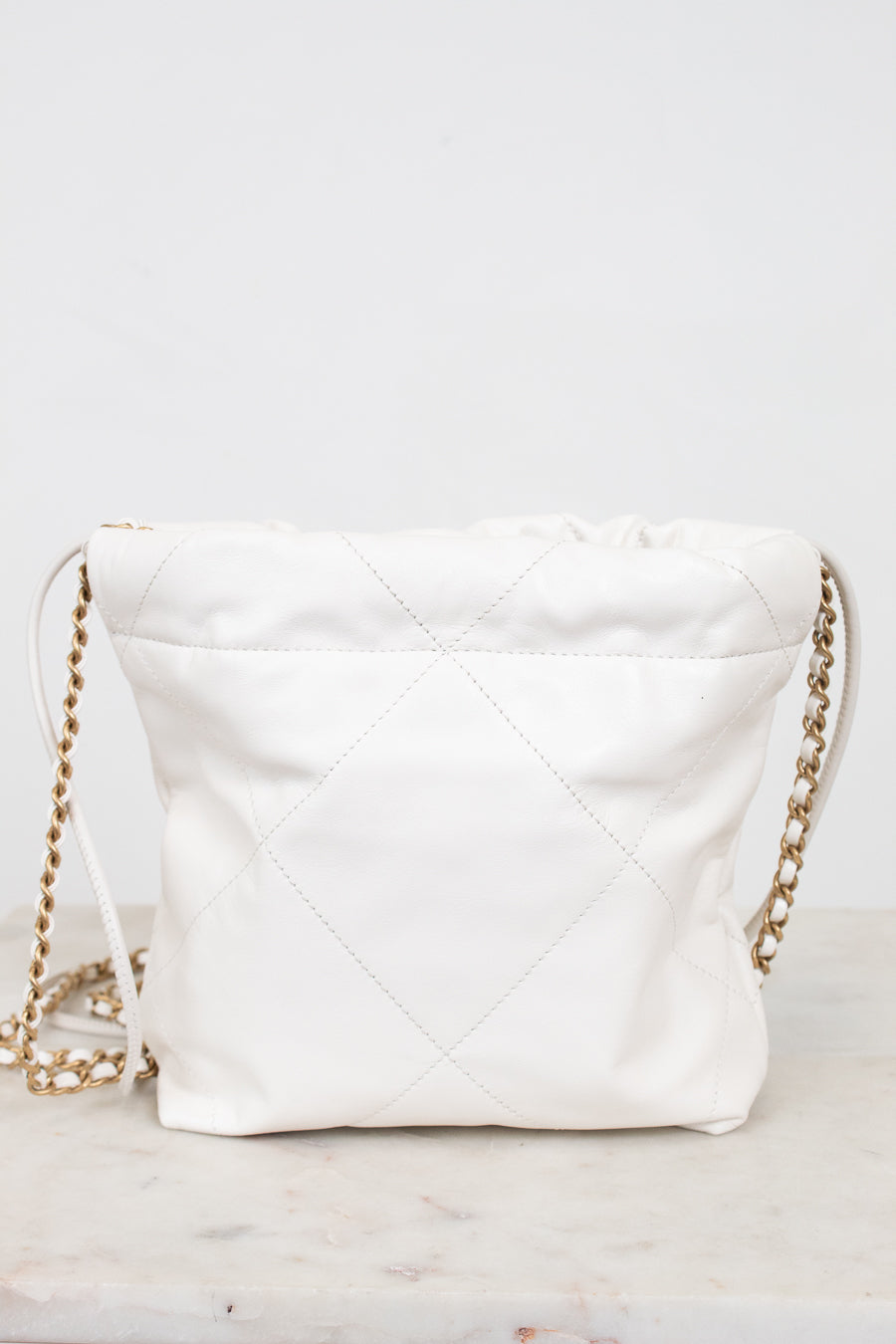 A white quilted leather shoulder bag with a gold chain strap, resting on a light marble surface against a white background.