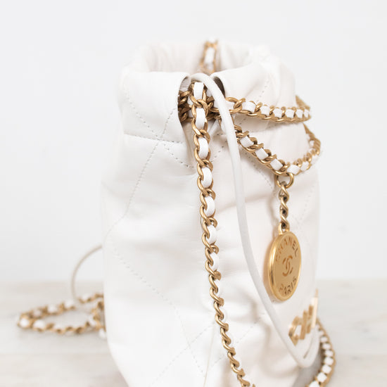 A white quilted leather bucket bag with gold-tone chain straps and a gold-tone CC logo medallion, resting on a light marble surface against a white background.