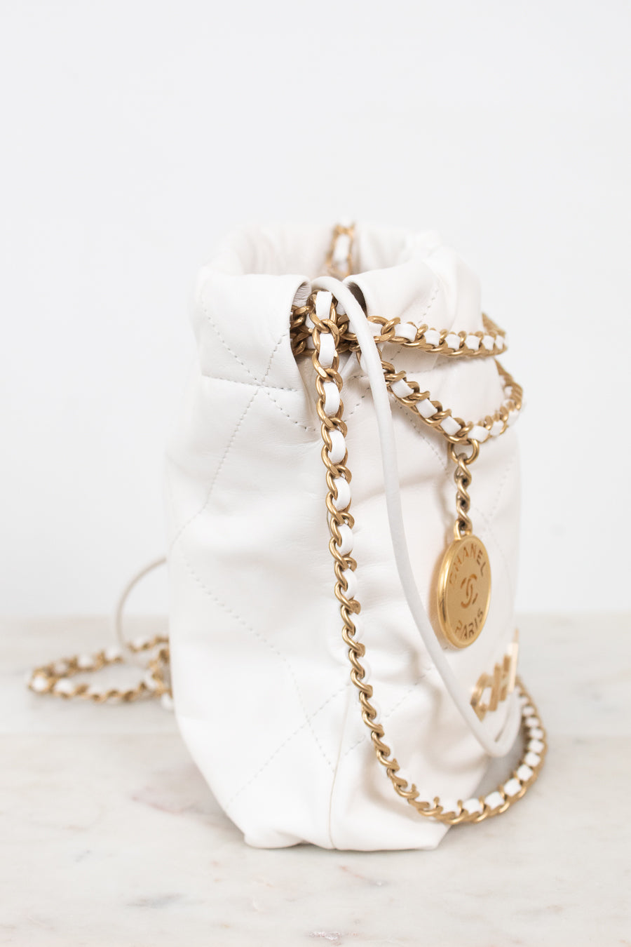 A white quilted leather bucket bag with gold-tone chain straps and a gold-tone CC logo medallion, resting on a light marble surface against a white background.