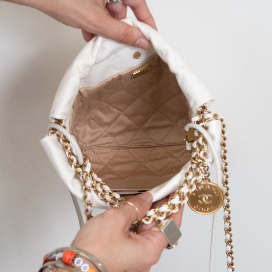 A close-up of a white Chanel bucket bag with gold chain straps, being held open by two hands. The bag features a white fabric exterior, a gold-tone chain strap, a gold interlocking CC charm, and a quilted beige/tan interior lining.