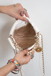 A close-up of a white Chanel bucket bag with gold chain straps, being held open by two hands. The bag features a white fabric exterior, a gold-tone chain strap, a gold interlocking CC charm, and a quilted beige/tan interior lining.