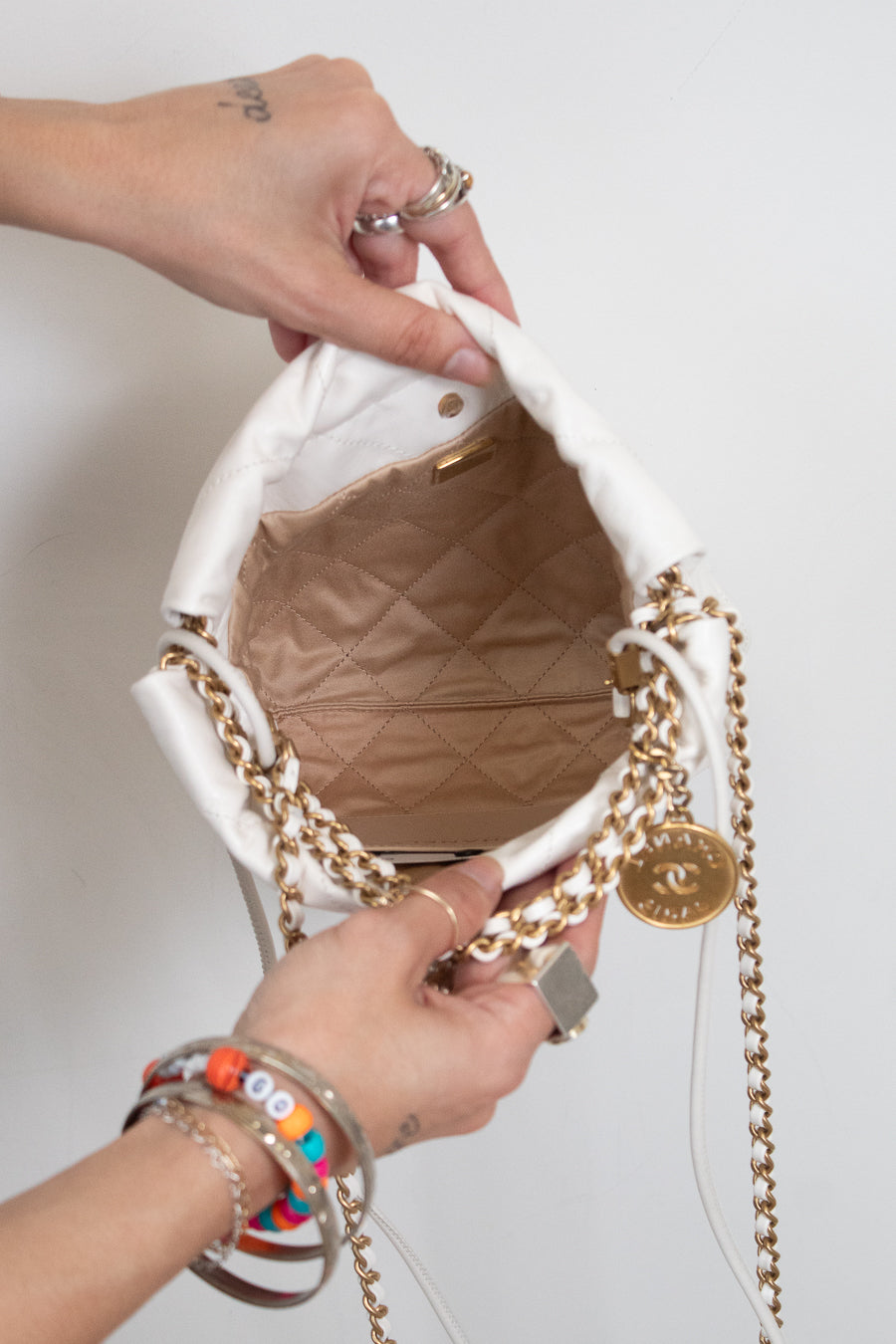 A close-up of a white Chanel bucket bag with gold chain straps, being held open by two hands. The bag features a white fabric exterior, a gold-tone chain strap, a gold interlocking CC charm, and a quilted beige/tan interior lining.