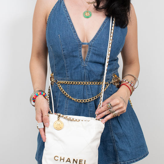 A model wearing a denim dress and holding a white Chanel crossbody bag with gold lettering and chain strap details, set against a plain light gray background.