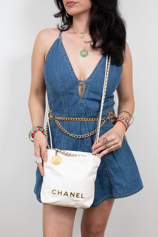 A model wearing a denim dress and holding a white Chanel crossbody bag with gold lettering and chain strap details, set against a plain light gray background.