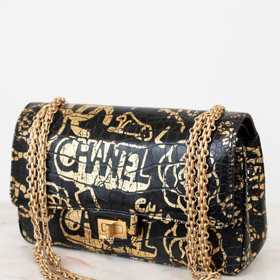 Black handbag with gold chain strap and graffiti-style text on a white background