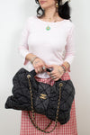 A person wearing a light pink long-sleeve top and a red-and-white gingham skirt holds a large black quilted Chanel handbag with gold-tone hardware and chain straps against a plain light background.