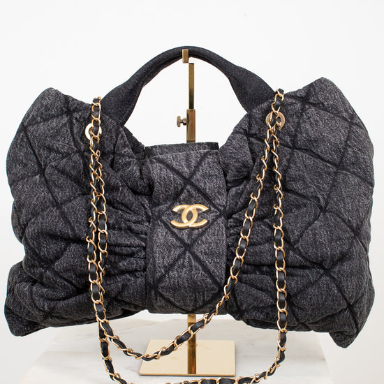 A gray quilted Chanel shoulder bag with gold-tone chain and handle, displayed on a stand over a white marble block against a white background.