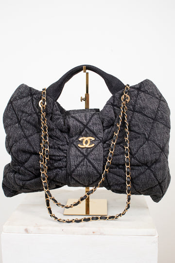 A gray quilted Chanel shoulder bag with gold-tone chain and handle, displayed on a stand over a white marble block against a white background.