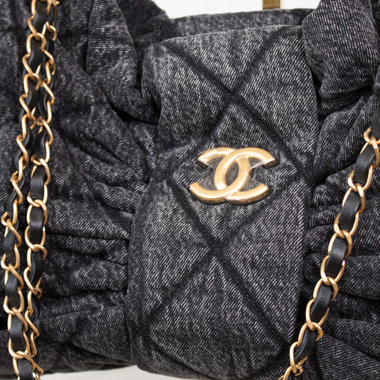 A close-up of a dark gray quilted Chanel shoulder bag with a gold-tone interlocking CC logo, featuring gathered detailing and a black-and-gold chain strap.