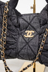 A close-up of a dark gray quilted Chanel shoulder bag with a gold-tone interlocking CC logo, featuring gathered detailing and a black-and-gold chain strap.